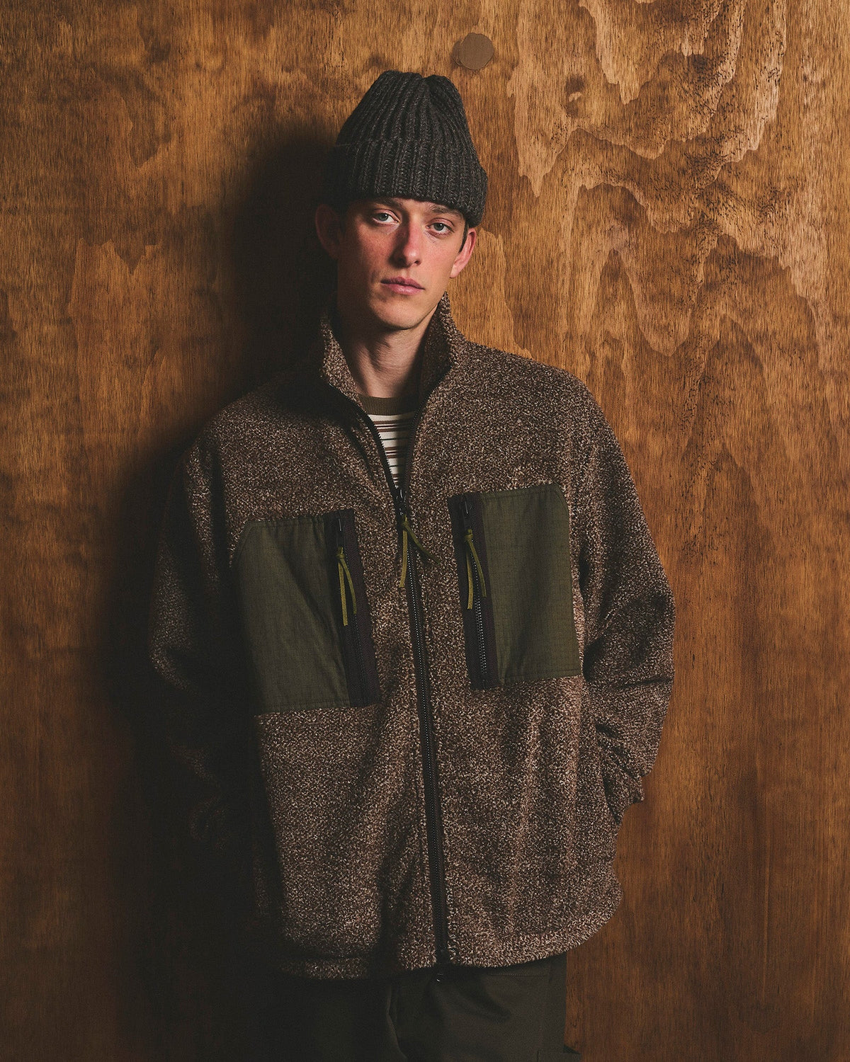 Man wearing a dark grey patchwork plaid chore jacket with olive corduroy collar over a black sweater and white t-shirt, paired with black trousers and a dark plaid baseball cap, standing against a wood-grain wall.