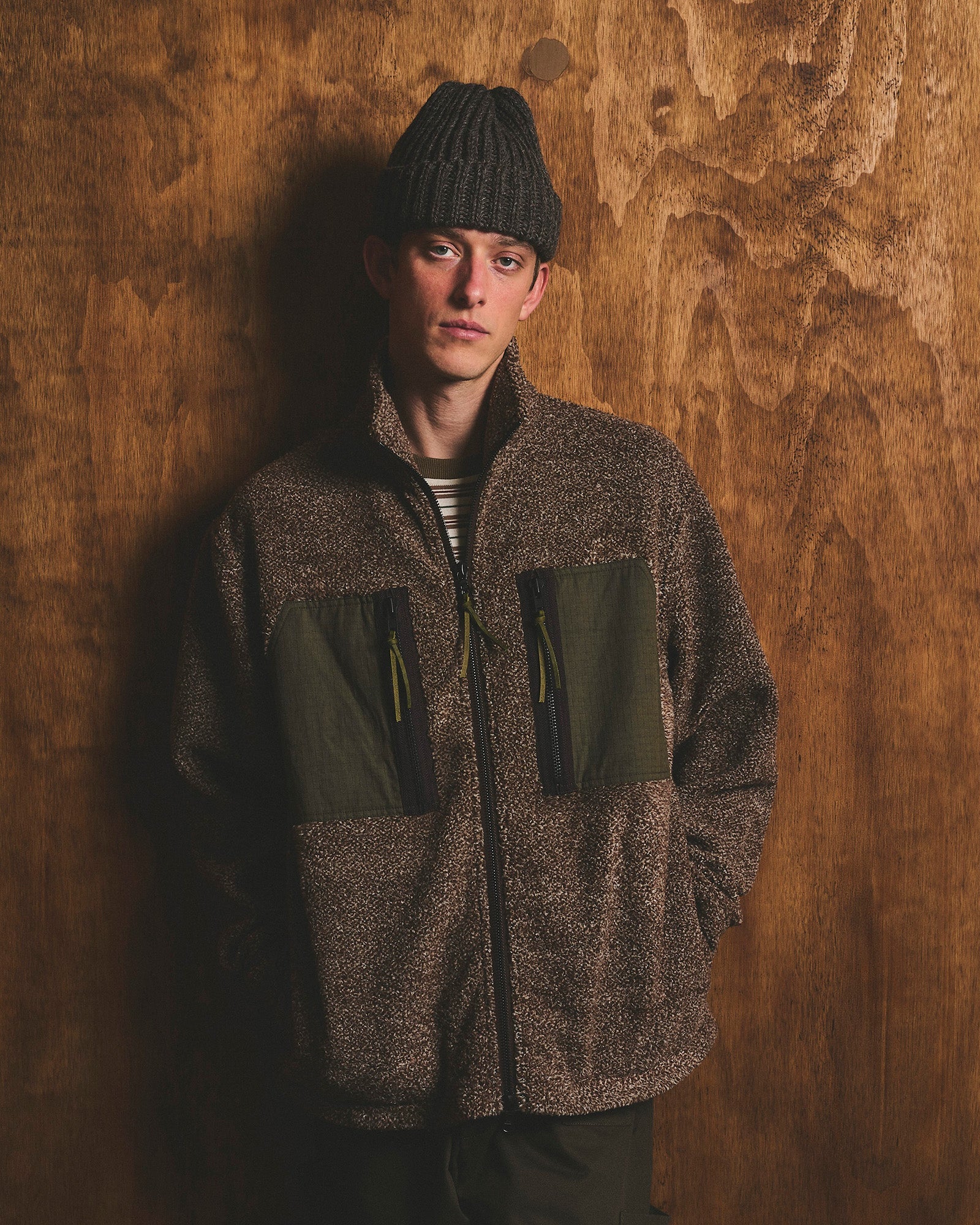 Man wearing a dark grey patchwork plaid chore jacket with olive corduroy collar over a black sweater and white t-shirt, paired with black trousers and a dark plaid baseball cap, standing against a wood-grain wall.