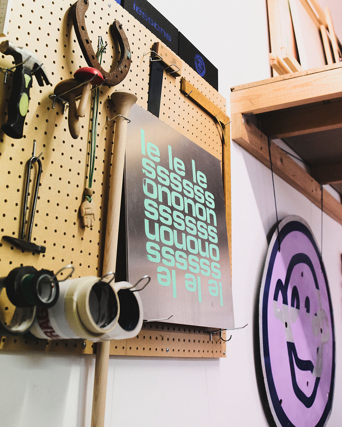 Hand tools, tape rolls and a horseshoe hang on a pegboard beside a metal sign spelling "lessons" in repeating mint-green letters, with a purple circular sign of a cartoon face on the wall nearby.